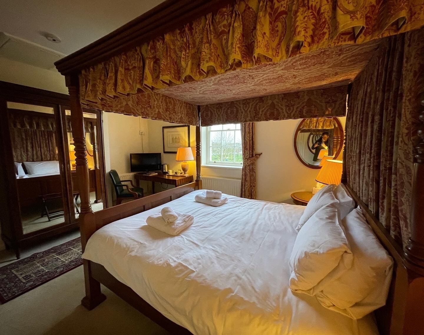 A four-poster bed with white linens and towels, a television, a lamp, and a mirror in a hotel room.