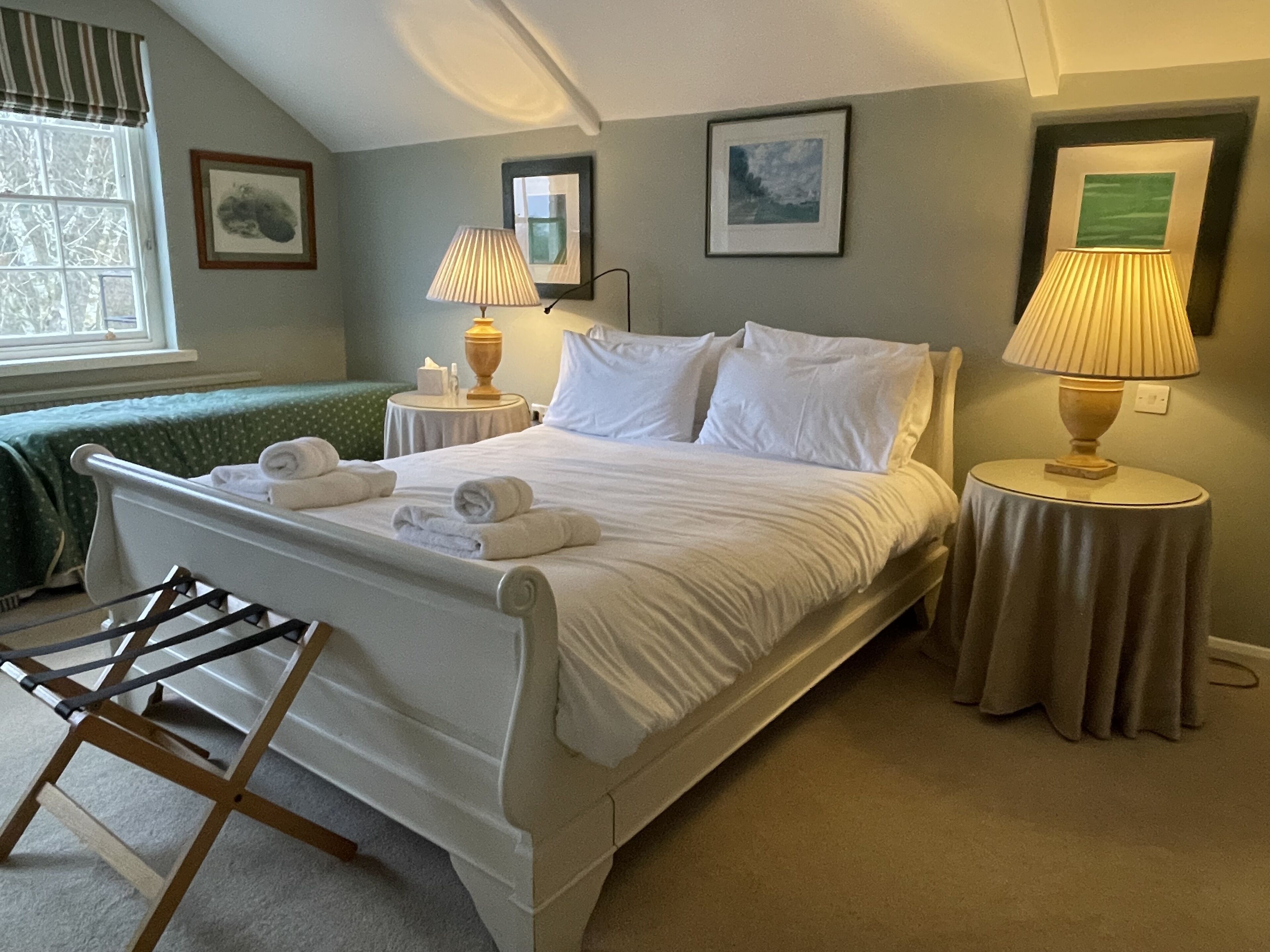 A neatly made bed with white linens and towels, two bedside lamps, a window with curtains, and a framed picture on the wall.