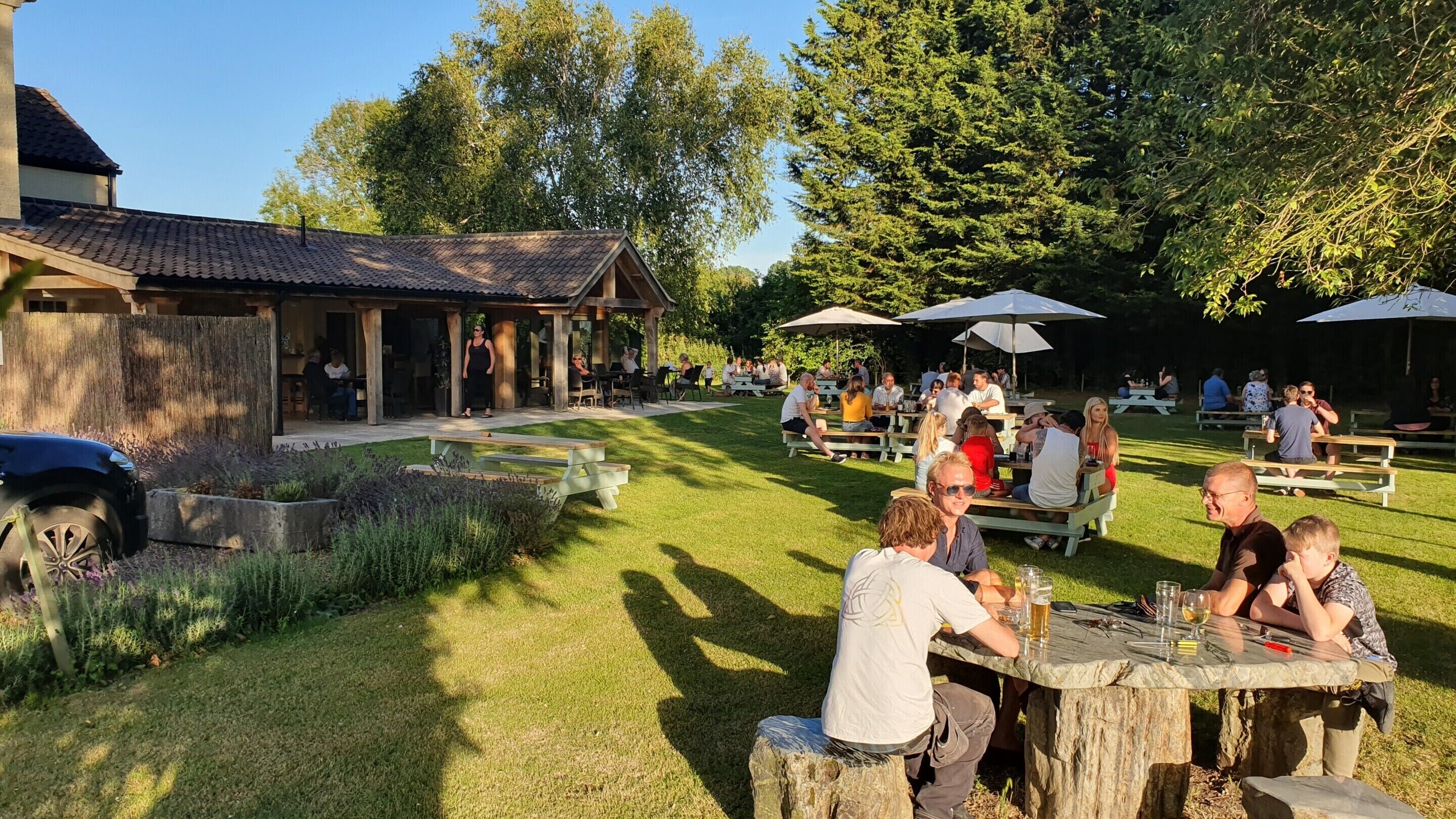 An outdoor dining area with wooden tables and benches, surrounded by a garden and trees.