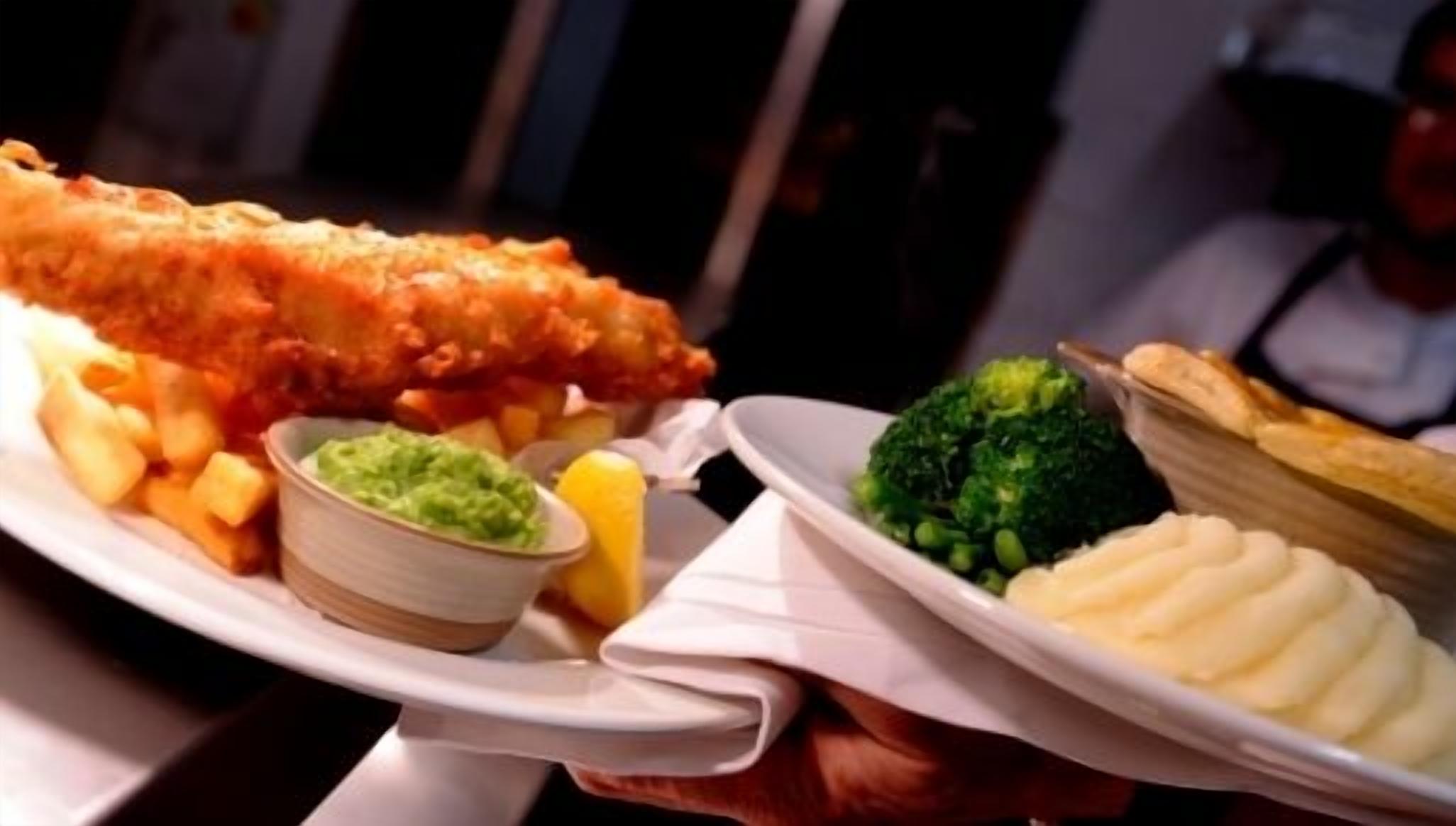 A plate of fish and chips with sides of mushy peas, chips, and a small bowl of tartar sauce.