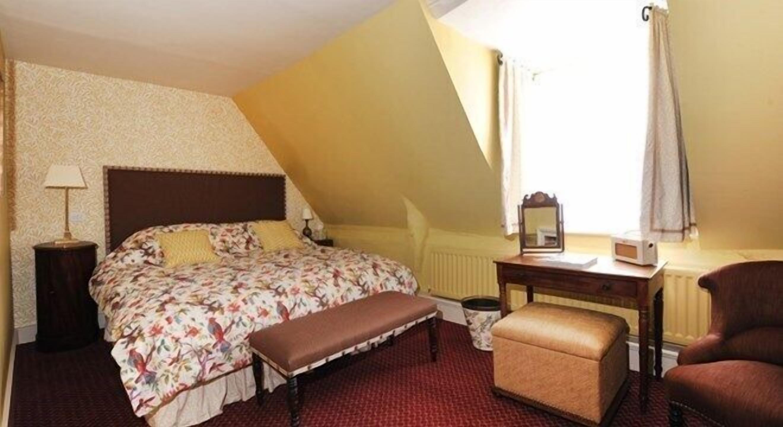 A bedroom with a bed, bench, desk, chair, lamp, and a mirror.