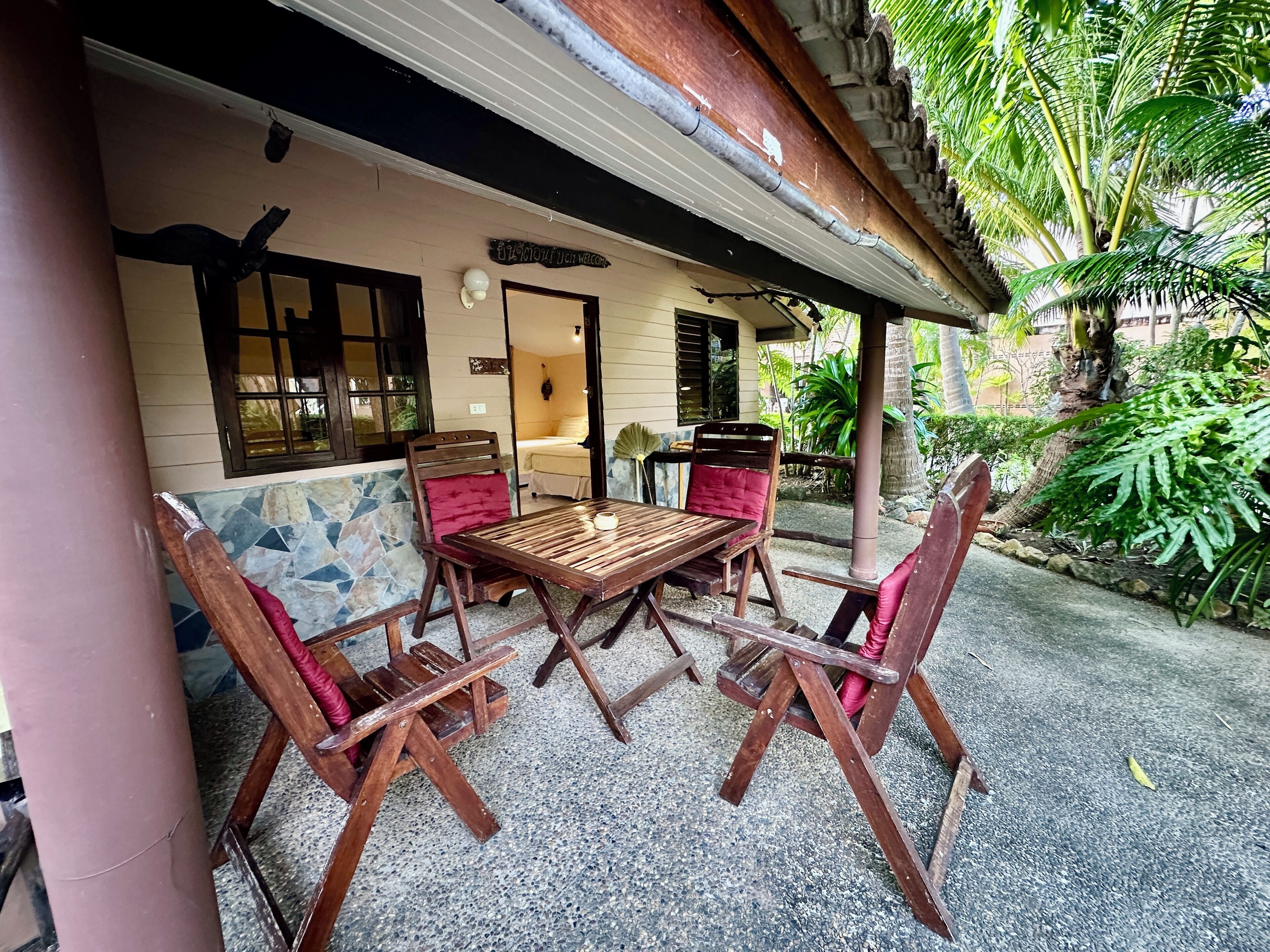 A wooden table and chairs set on a veranda with a tiled floor, a sign reading "Welcome", and a view of a room with a bed and window.