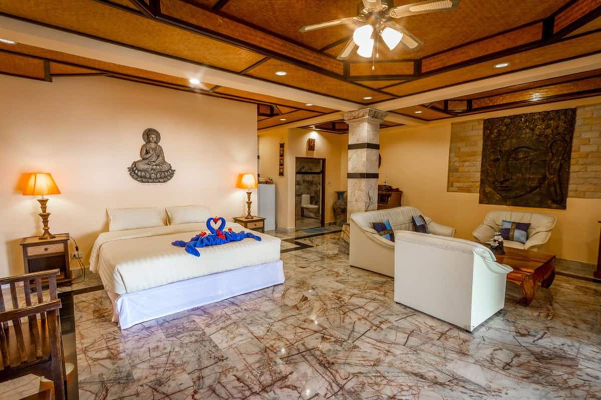 A hotel room with a bed, two chairs, a sofa, a ceiling fan, and a Buddha statue on the wall.