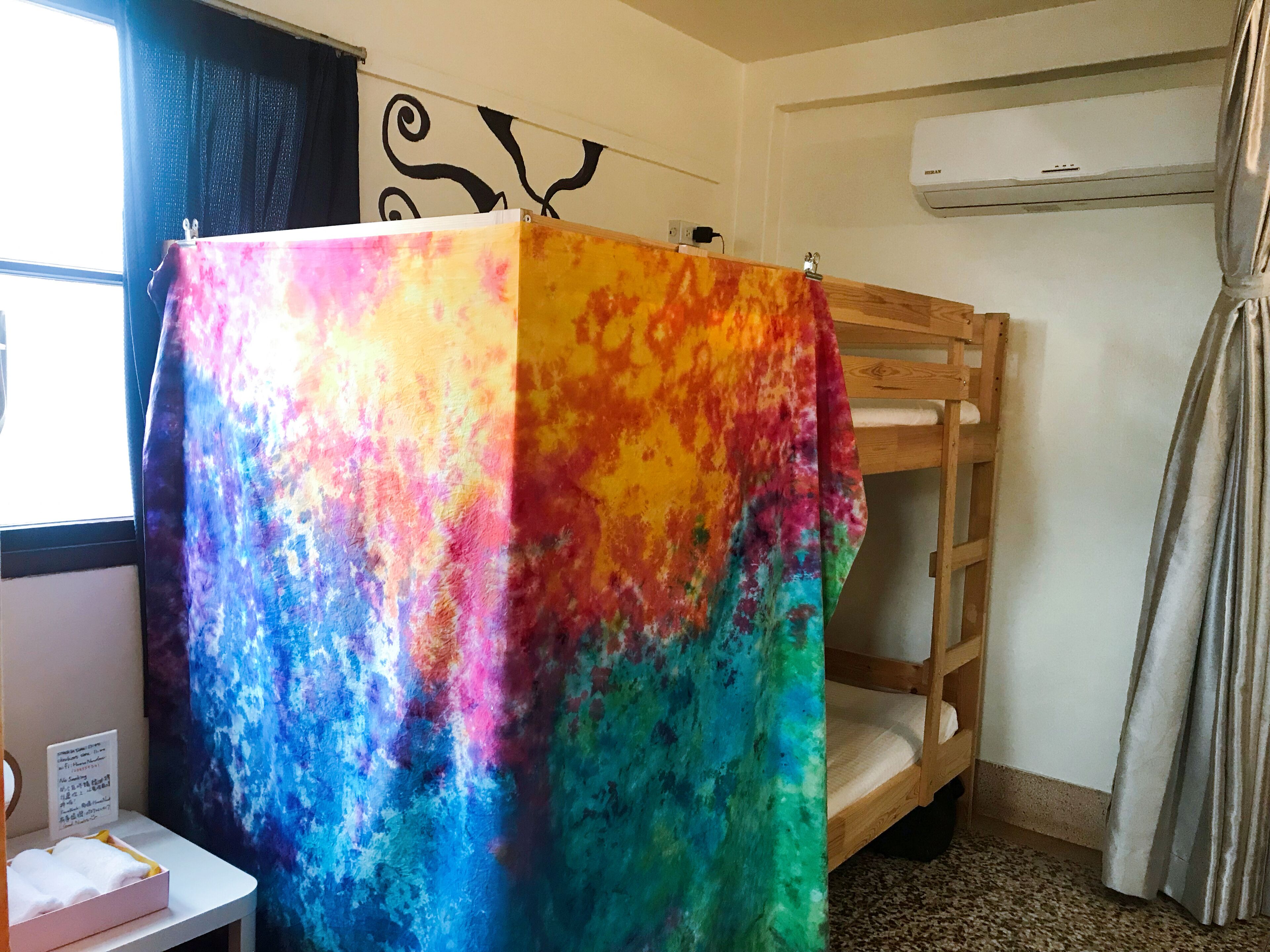 Una habitación con una cama litera, una manta colorida con estampado de técnica tie-dye, un escritorio pequeño con una silla, una ventana con cortinas y un equipo de aire acondicionado.