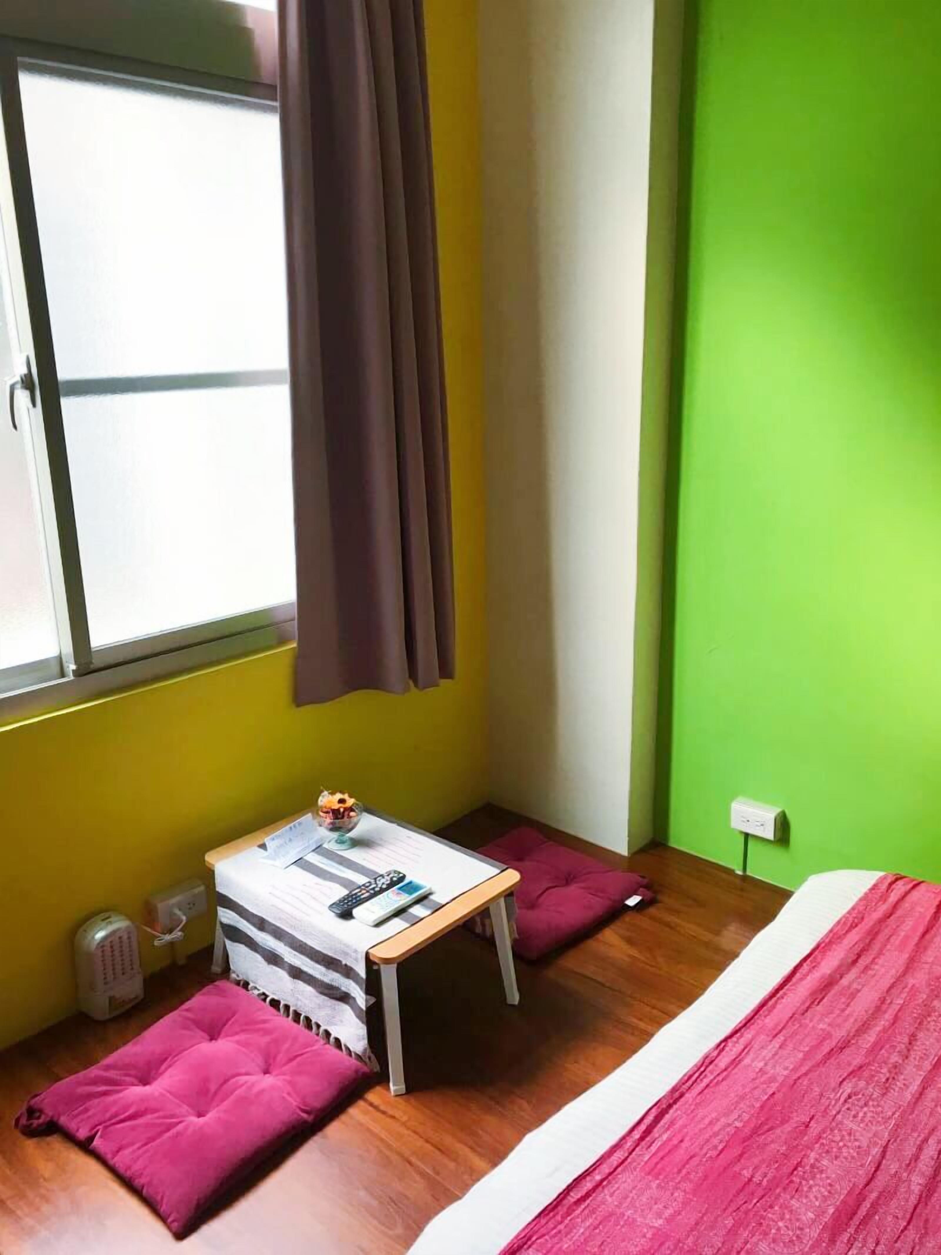 A room with a bed, a small table with a remote control, a window with curtains, and a green wall.