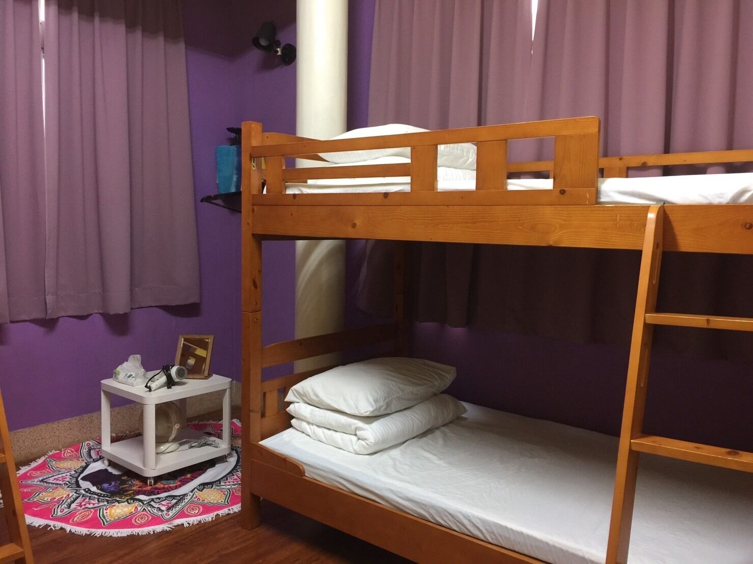 A single wooden bunk bed with a white mattress and pillows, a small white nightstand with a lamp, and a colorful rug.