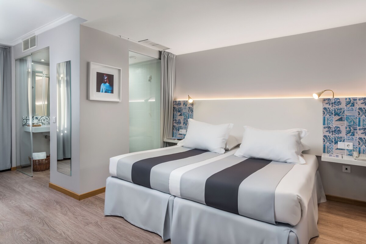 A hotel room with a large bed, a mirror, a picture on the wall, and a bathroom visible through a glass door.