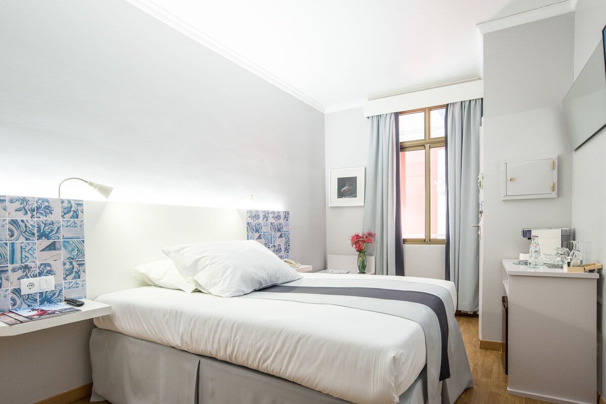 A hotel room with a bed, bedside table, lamp, and a window with curtains.