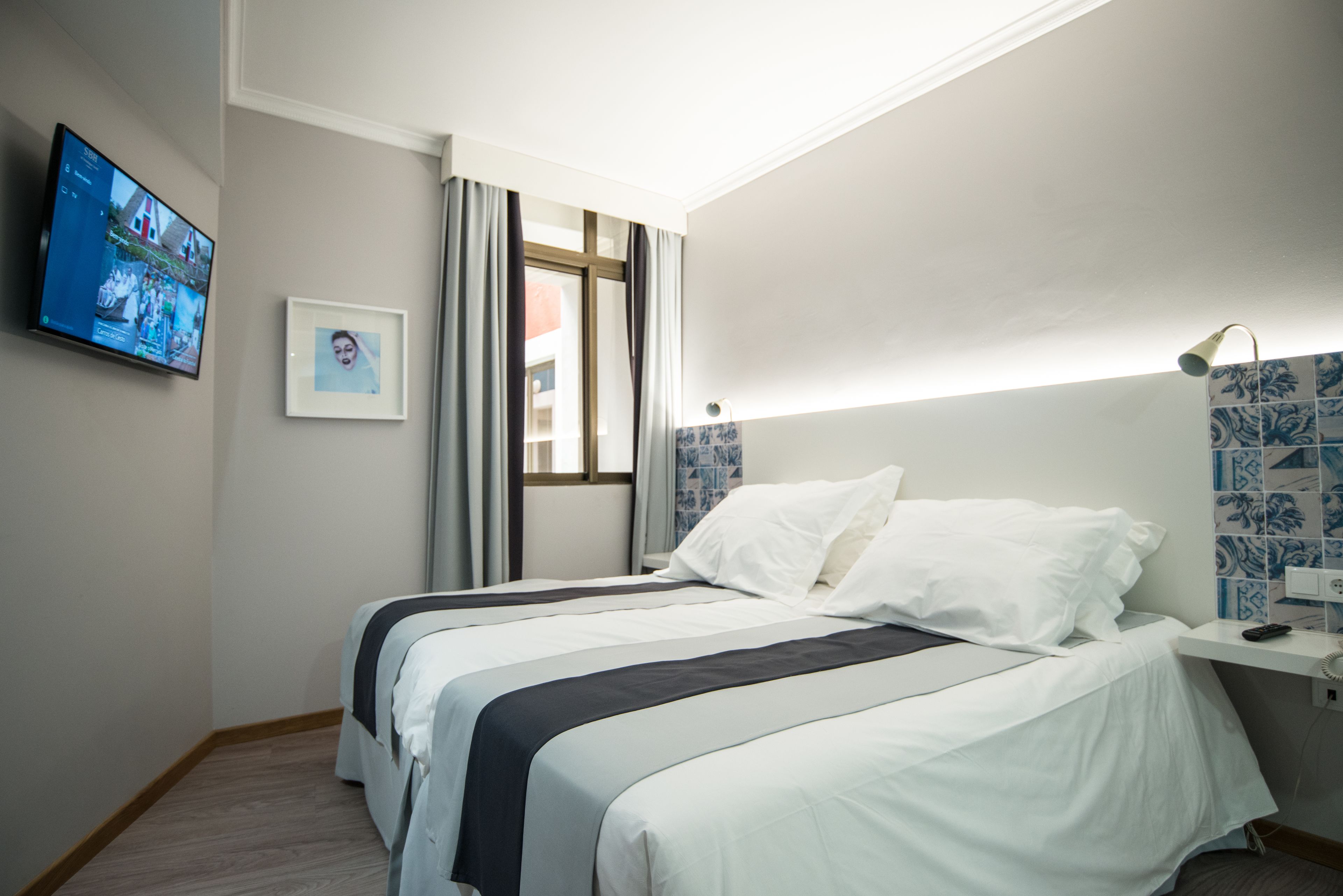 Habitación de hotel con cama, televisor montado en la pared, un cuadro enmarco y una ventana con cortinas.