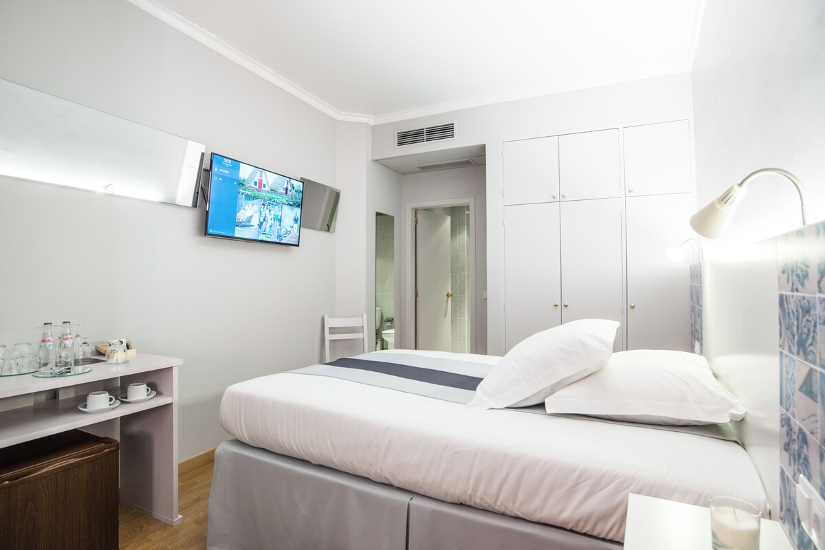 A modern hotel room with a bed, a TV mounted on the wall, a desk with a chair, and a wardrobe.