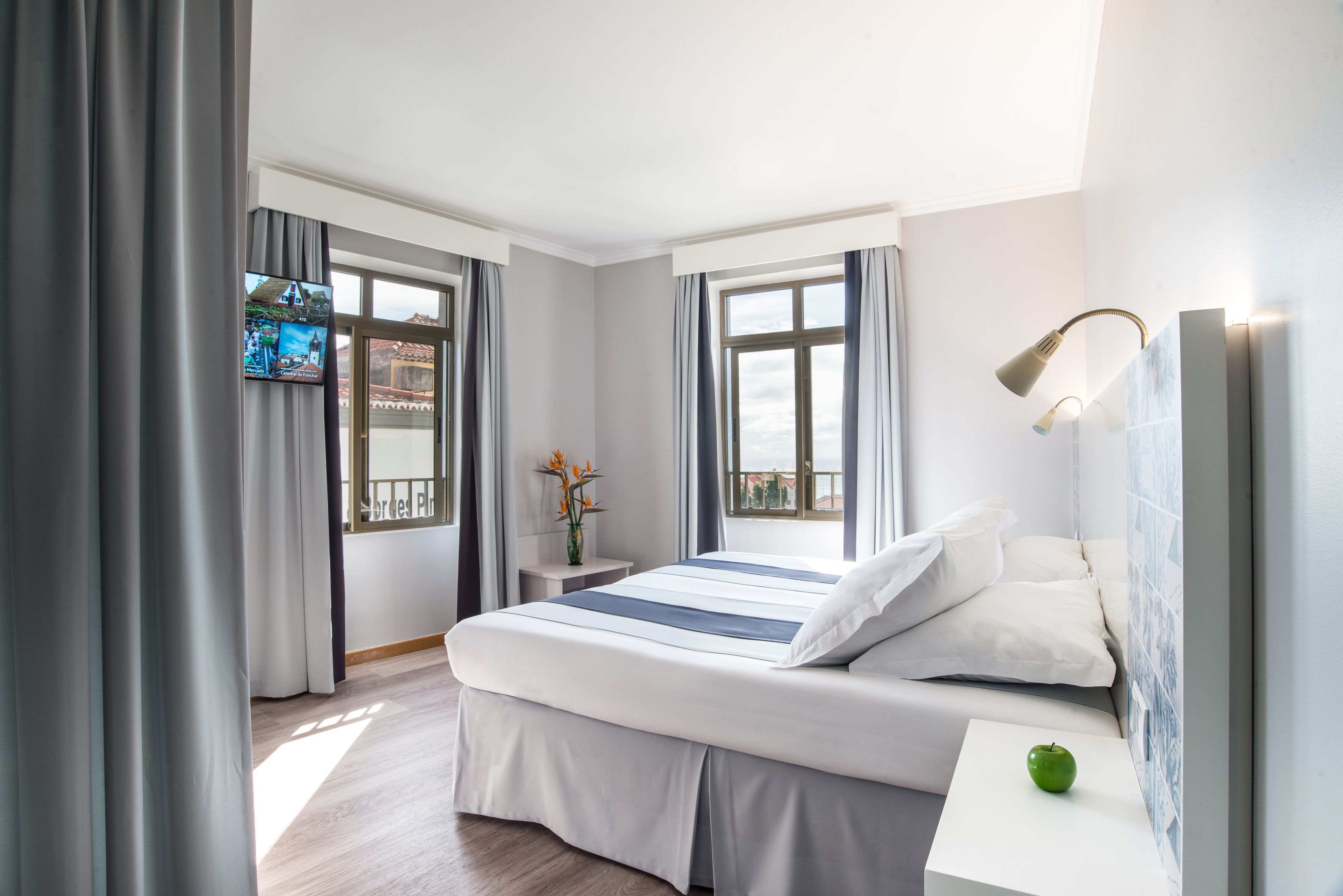 Una habitación de hotel moderna con una cama grande, una mesita de noche pequeña, un televisor montado en la pared y una ventana con cortinas.