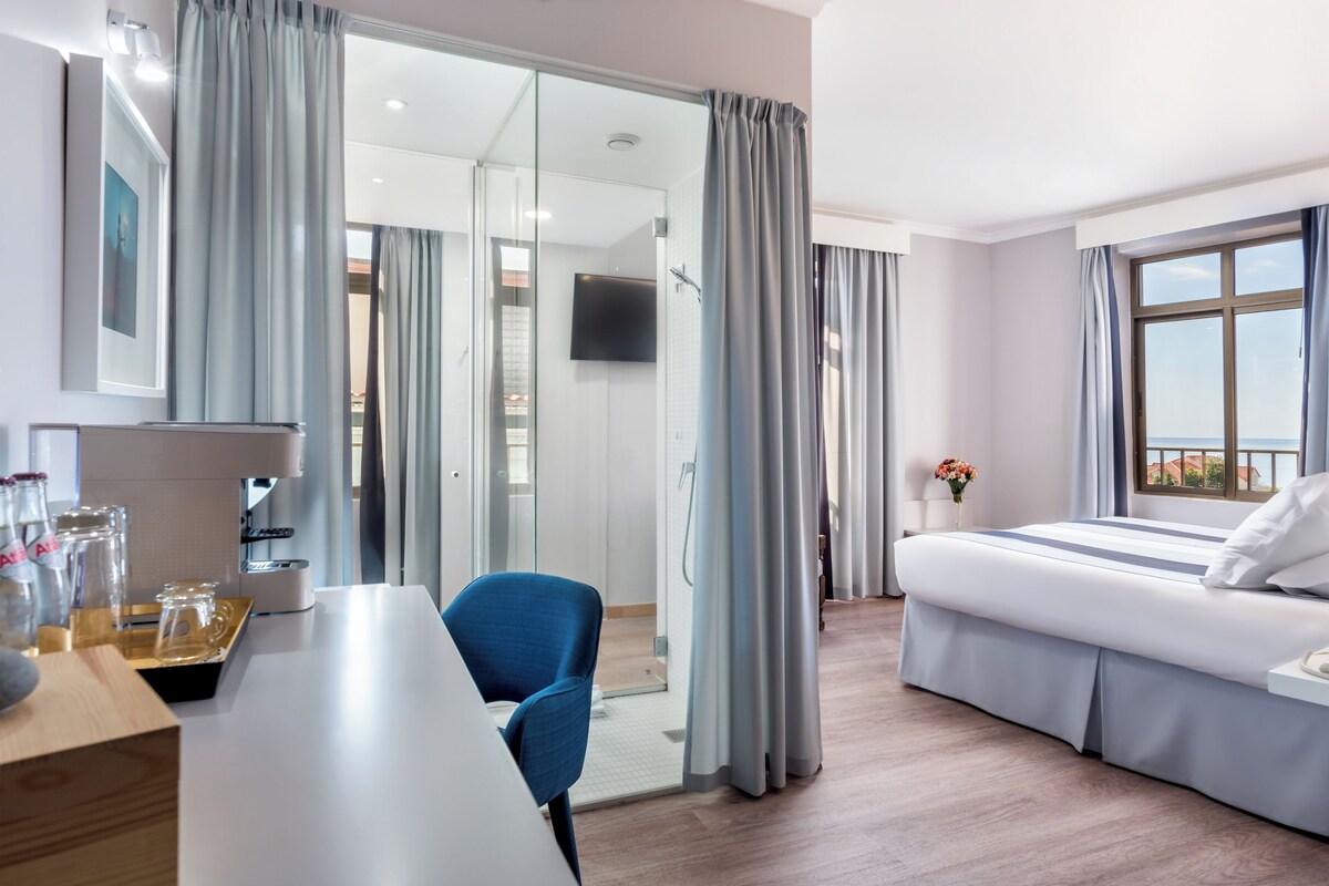 A modern hotel room with a bed, a desk, a chair, and a shower area.