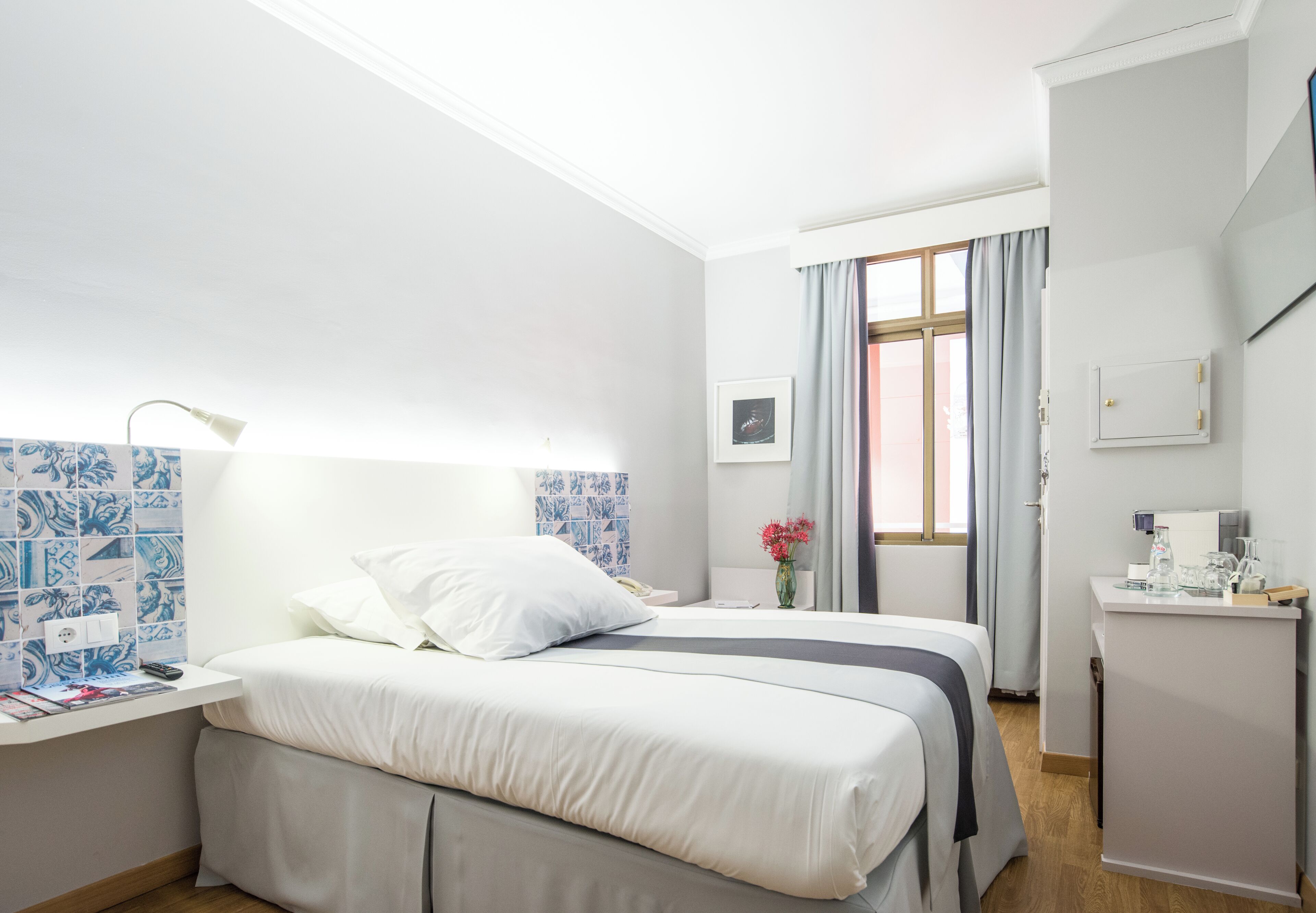 A hotel room with a bed, a desk with a lamp, a small table with a coffee maker, and a window with curtains.