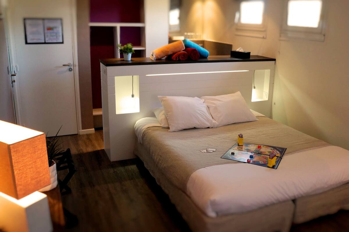 Una habitación de hotel con una cama, mesita de noche, lámpara y una pequeña mesa con un juego de mesa.