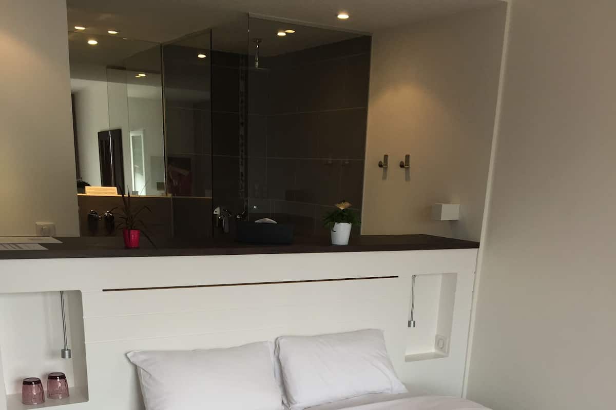 Un dormitorio moderno con una cama, ropa de cama blanca y almohadas rojas. Hay un estante pequeño con adornos y un espejo sobre la cama.