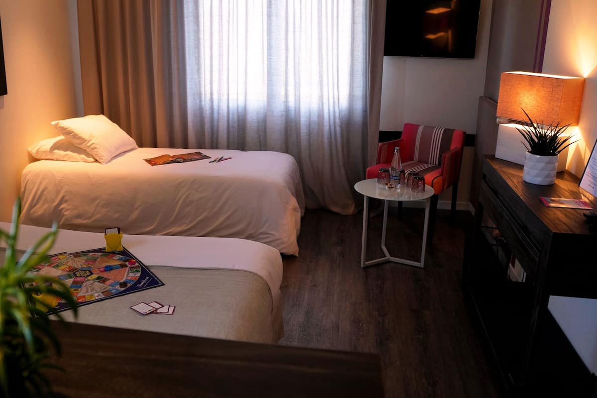 Habitación de hotel con dos camas, una mesita redonda, una silla roja, una lámpara y una planta en maceta.