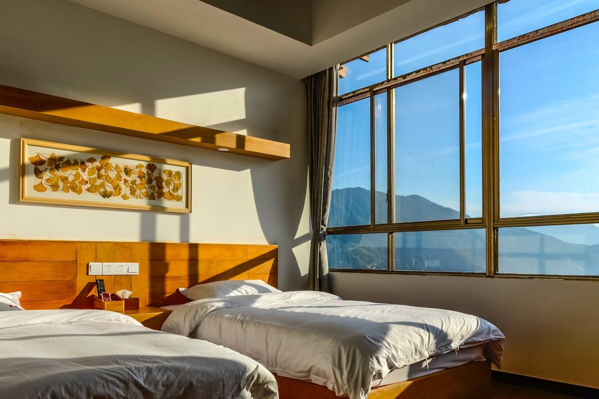 A hotel room with two beds, a wooden headboard, a large window with a mountain view, and a framed artwork on the wall.