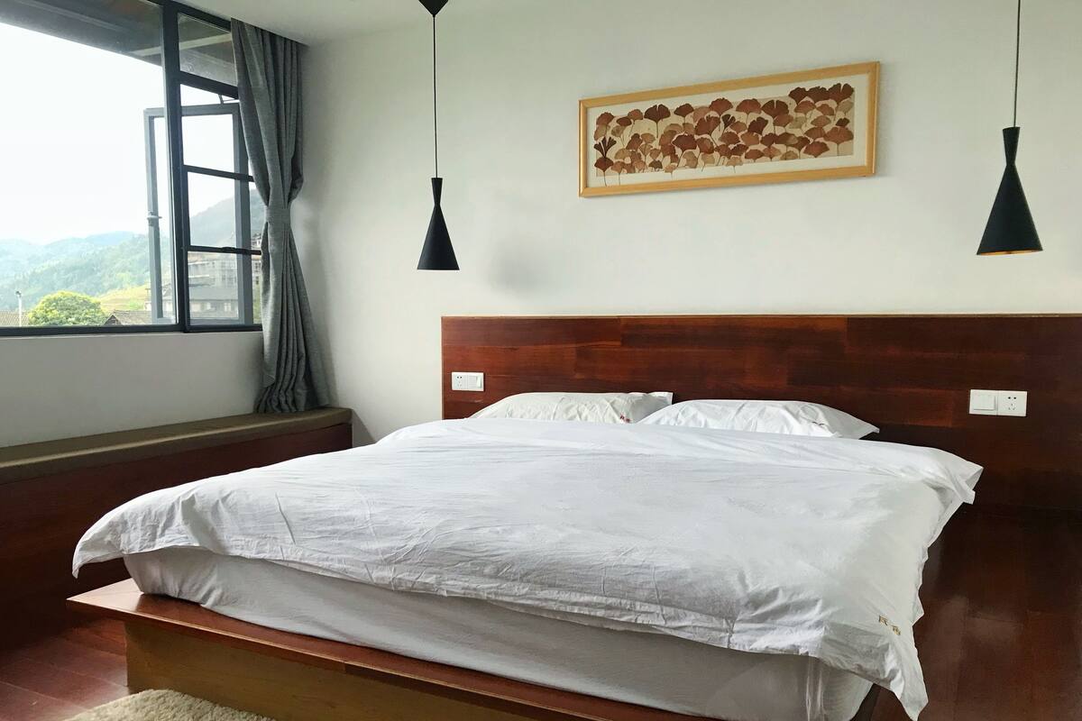 A neatly made bed with white linens, a wooden headboard, and a view of greenery through a window with curtains.