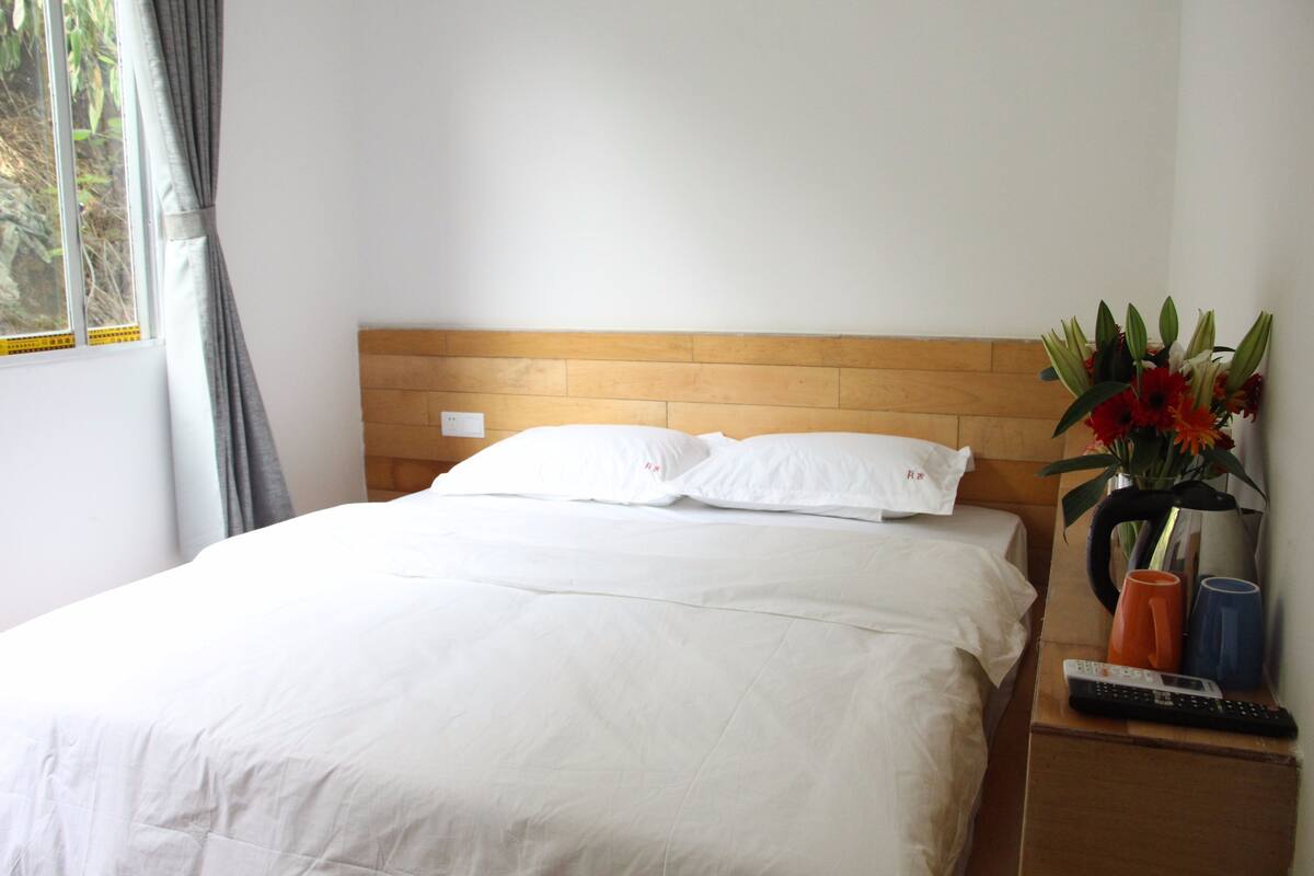 A neatly made bed with white linens, a wooden headboard, a nightstand with a remote control, a vase with flowers, and a window with curtains.