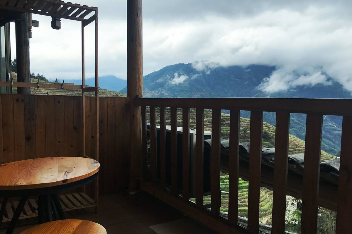 A wooden balcony with a table and chairs, overlooking terraced fields and mountains.