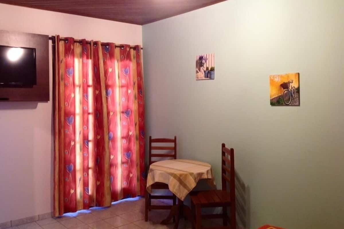 A small room with a TV mounted on the wall, a table with a tablecloth, two chairs, and a window with curtains.