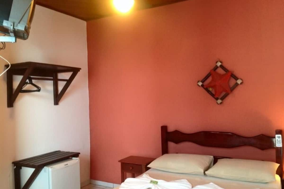 A bedroom with a bed, a small bedside table, a hanging light fixture, and a wall-mounted shelf.