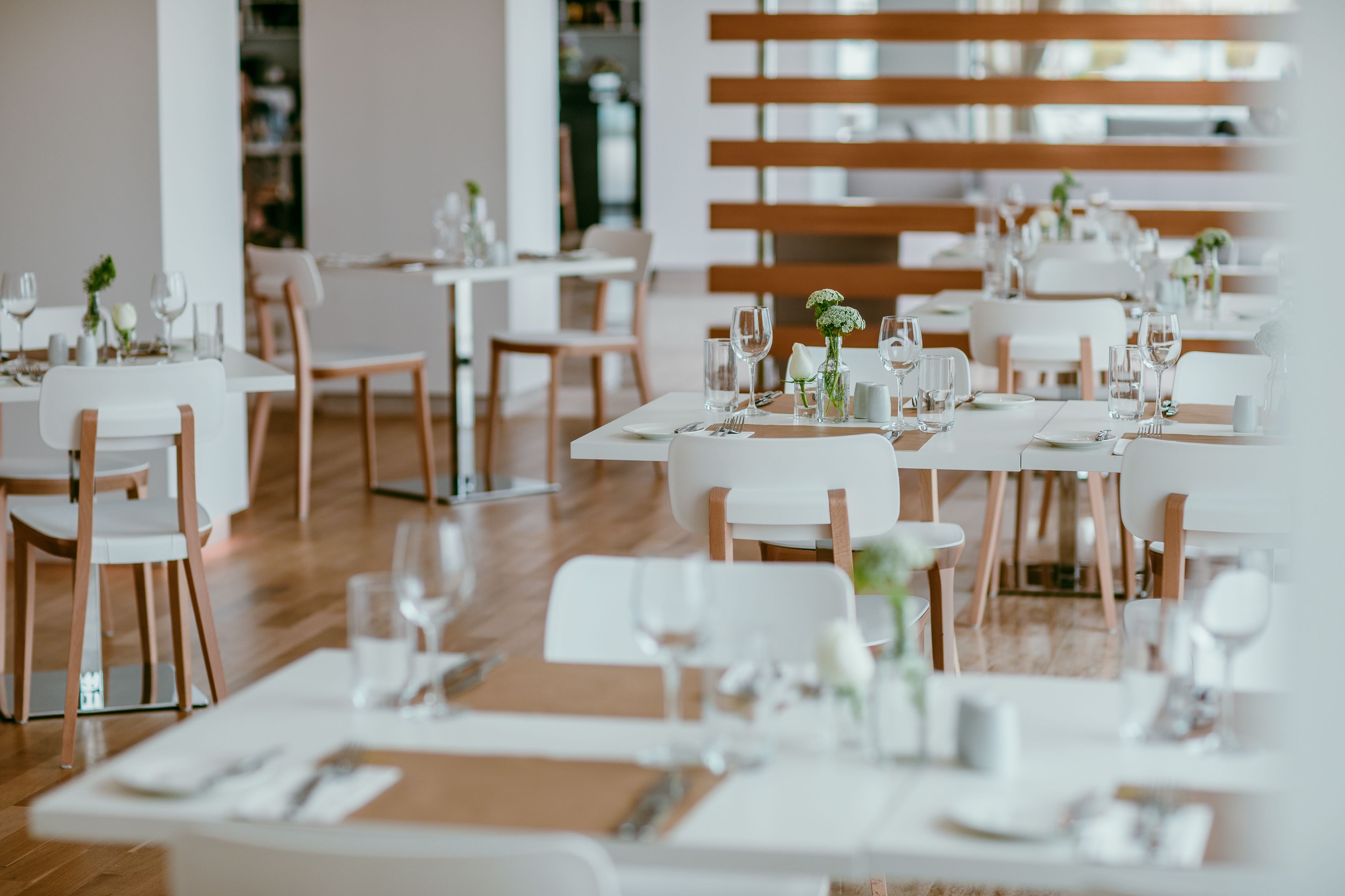 Un restaurante con mesas y sillas blancas, ordenadamente dispuestas con copas de vino y cubiertos.
