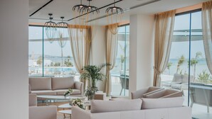 A modern hotel lobby with a large seating area, a glass wall offering a view of the sea, and a potted plant. - Lebay Beach Hotel (Oroklini)