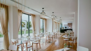 A modern restaurant with white tables and chairs, large windows, and a kitchen in the background. - Lebay Beach Hotel (Oroklini)