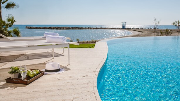 A poolside area with a clear view of the ocean, a lounge chair, a tray with apples and a drink, and a hat on the deck. - Lebay Beach Hotel (Oroklini)