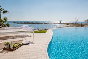 A poolside area with a clear view of the ocean, a lounge chair, a tray with apples and a drink, and a hat on the deck. - Lebay Beach Hotel (Oroklini)