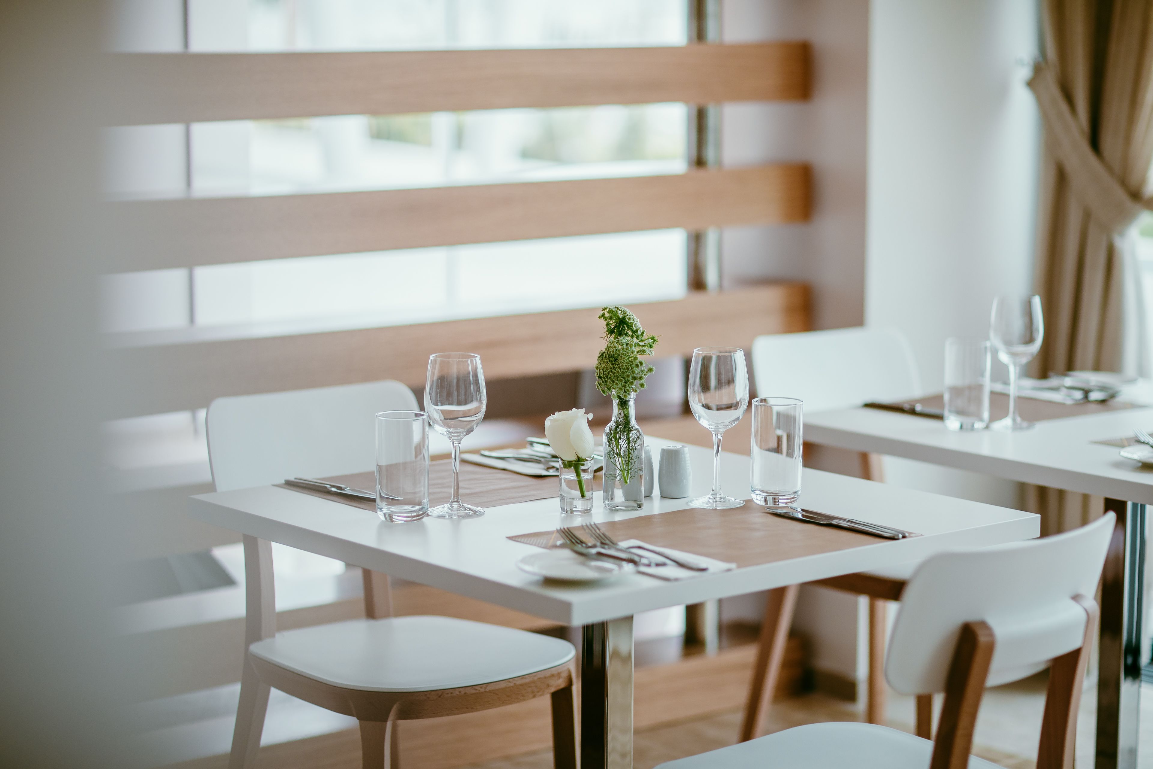 A dining area with white tables and chairs, set with glassware and a small vase with a flower.