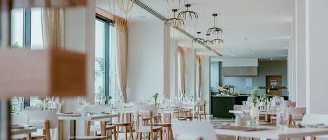 Un comedor moderno con mesas y sillas blancas, iluminación colgante y ventanas amplias.