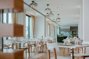 Un comedor moderno con mesas y sillas blancas, iluminación colgante y ventanas amplias.