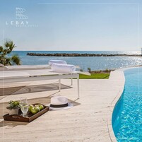 Un espace aménagé au bord de la piscine, avec vue sur l’océan, comprenant un fauteuil de relaxation, une table avec un plateau de pommes et des verres, et un chapeau.