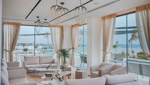A modern lounge with a sofa, armchairs, and a coffee table. Large windows offer a view of the ocean and a pool. - Lebay Beach Hotel (Oroklini)