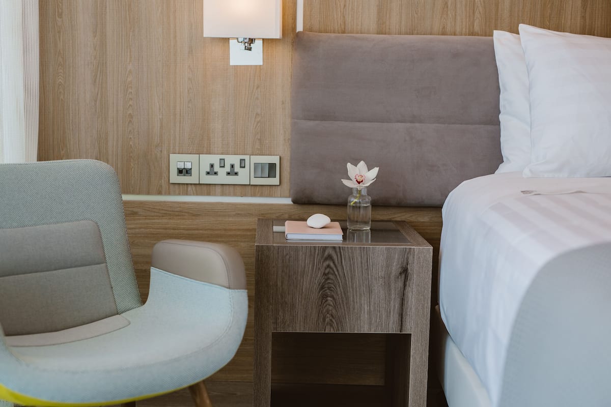 A hotel room with a wooden headboard, a bedside table with a vase of flowers, a chair, and a bed with white bedding.