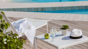 A poolside lounge with a white chair, a table with a green apple, two glasses of water, a small potted plant, and a white hat.