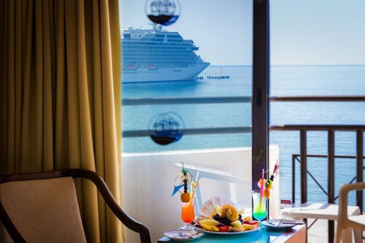 A room with a view of a cruise ship on the water, a table with fruit and drinks, and a window with curtains.