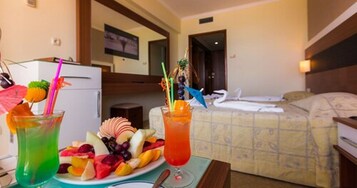 Habitación de hotel con una cama, una mesita de noche con una bandeja de frutas y bebidas, y un televisor al fondo.