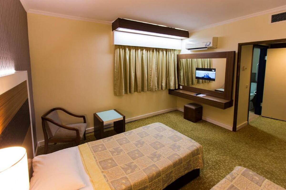 A hotel room with a bed, a chair, a small table, a TV mounted on the wall, and an air conditioning unit.