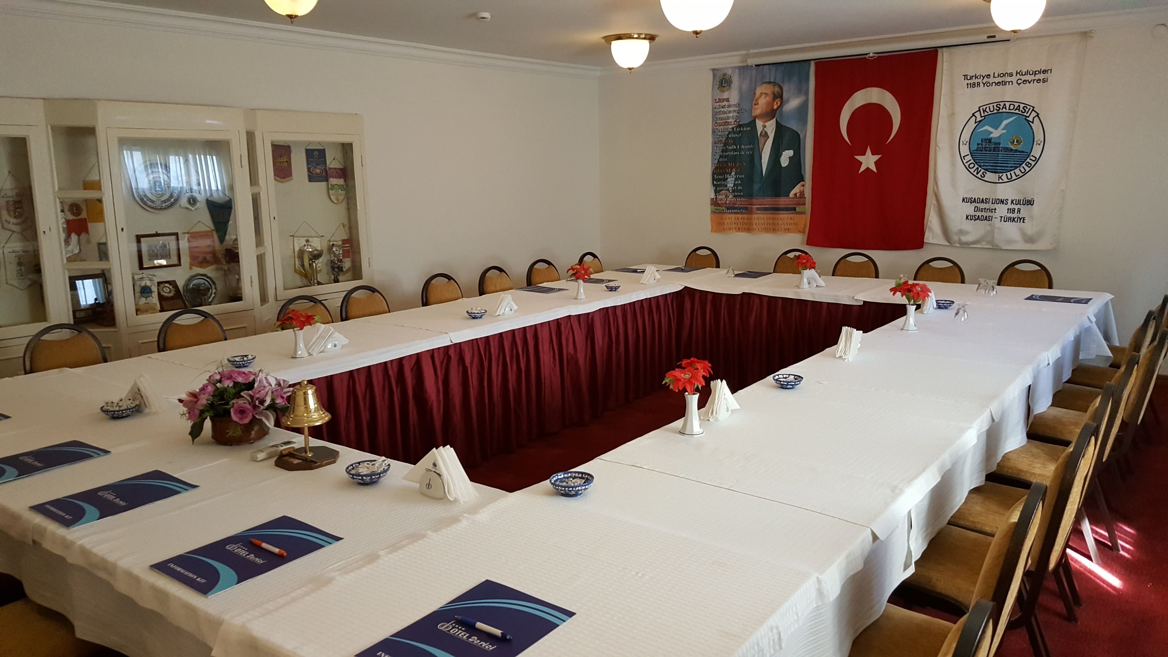 Uma sala de conferências com uma mesa comprida, cadeiras e uma divisória de cortina vermelha. Há bandeiras e um banner na parede.