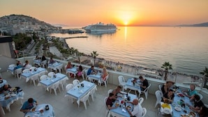 A rooftop dining area with tables and chairs overlooking a sunset and a cruise ship. - Derici Hotel (Kusadasi)