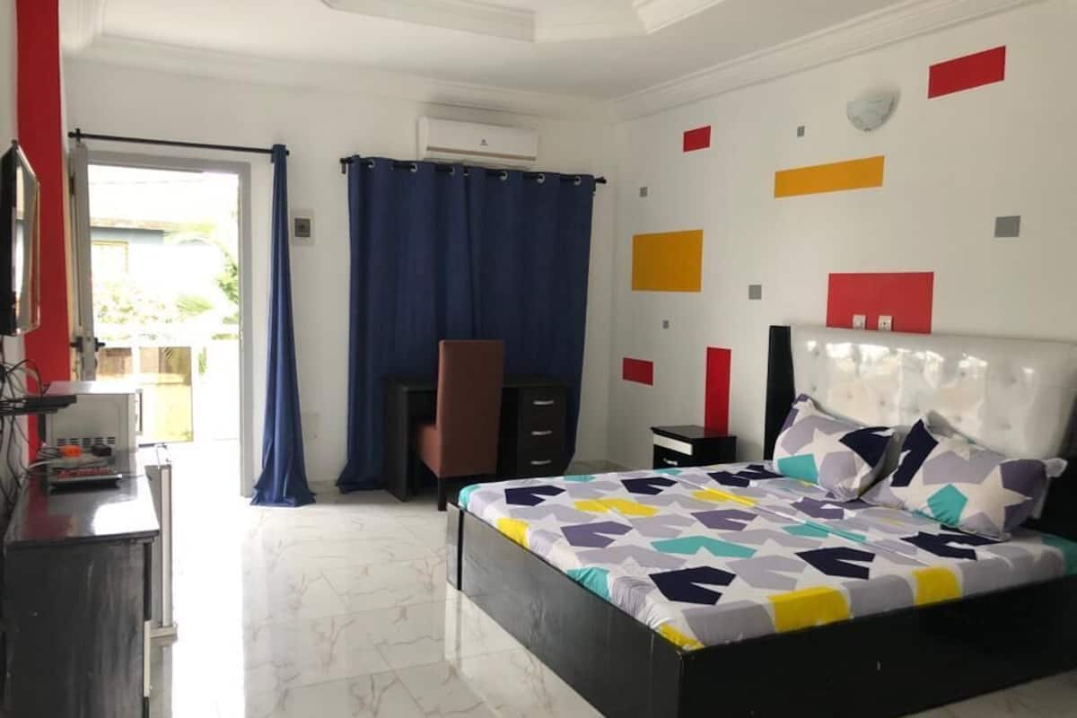 Un dormitorio con cama, escritorio y silla. Hay una ventana con cortinas, aire acondicionado montado en la pared y una puerta que da al exterior.