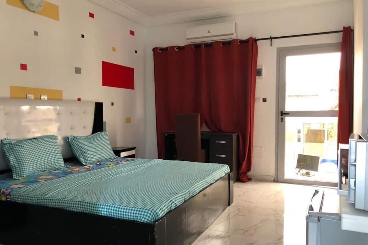 Un dormitorio con cama, cómoda, escritorio y ventana con cortinas rojas.