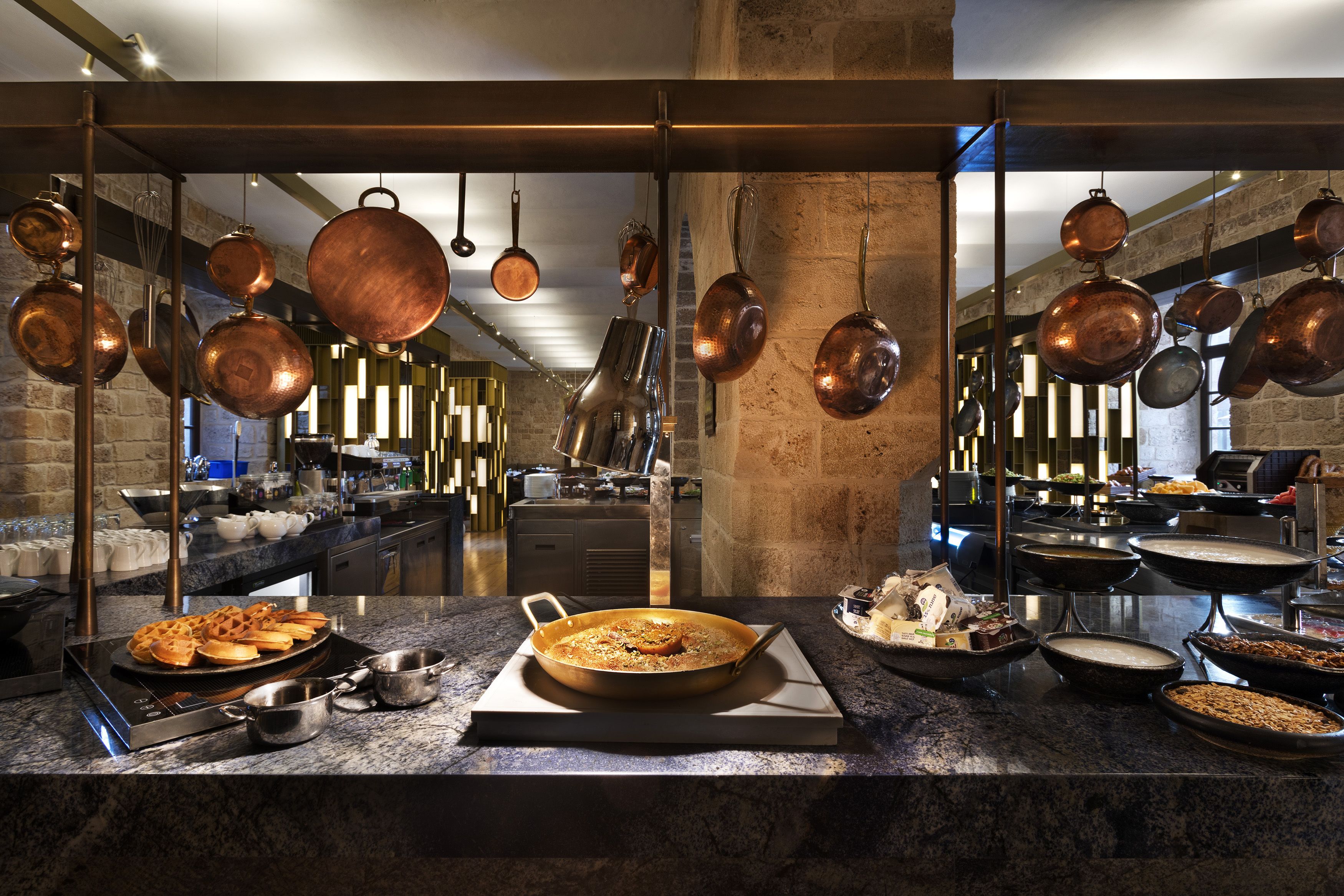 A kitchen with copper pots hanging from the ceiling, a marble countertop with various dishes, and a stone wall in the background.