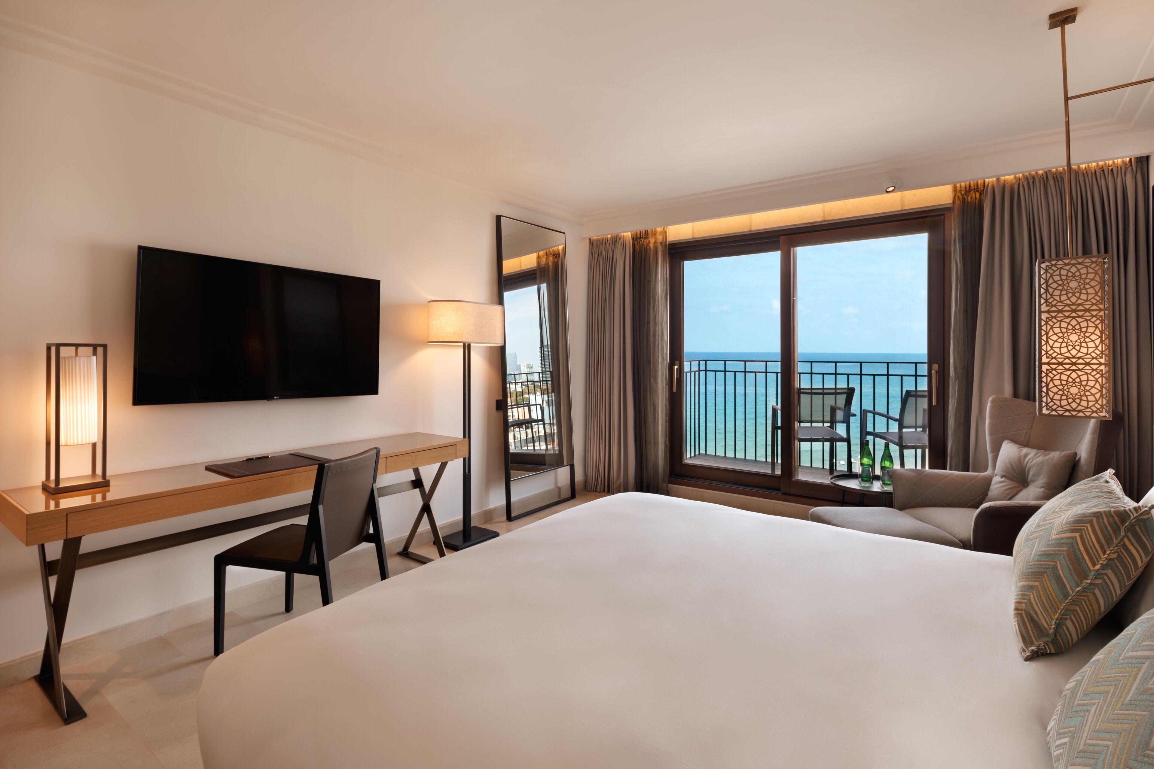 Habitación de hotel con una cama grande, un escritorio con silla, televisión montada en la pared y un balcón con vistas al mar.