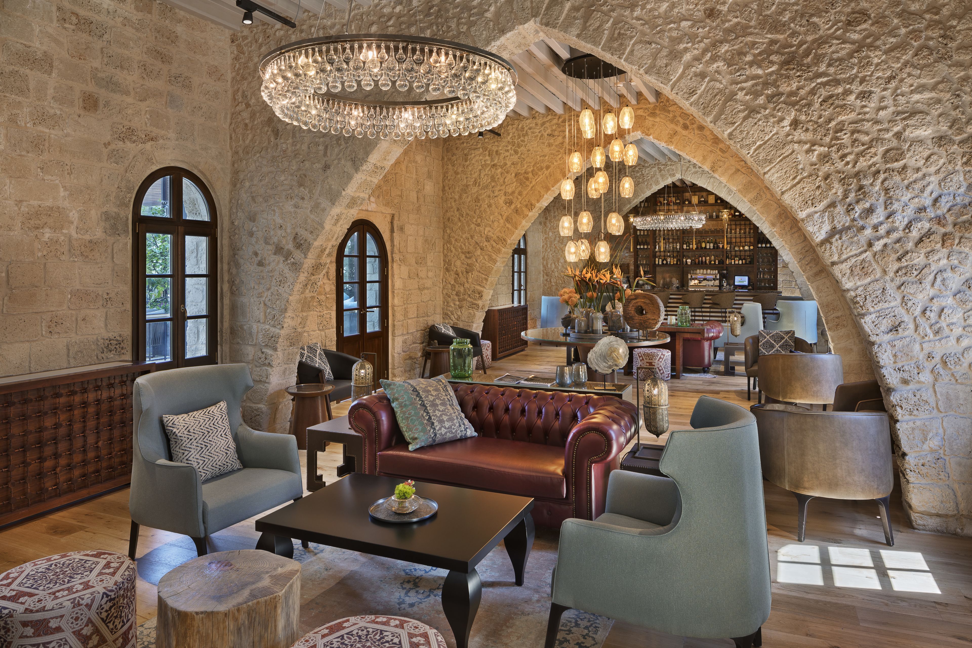 A spacious lounge area with leather sofas, armchairs, and a wooden coffee table. A chandelier hangs from the ceiling, and the room features exposed stone walls and large windows.