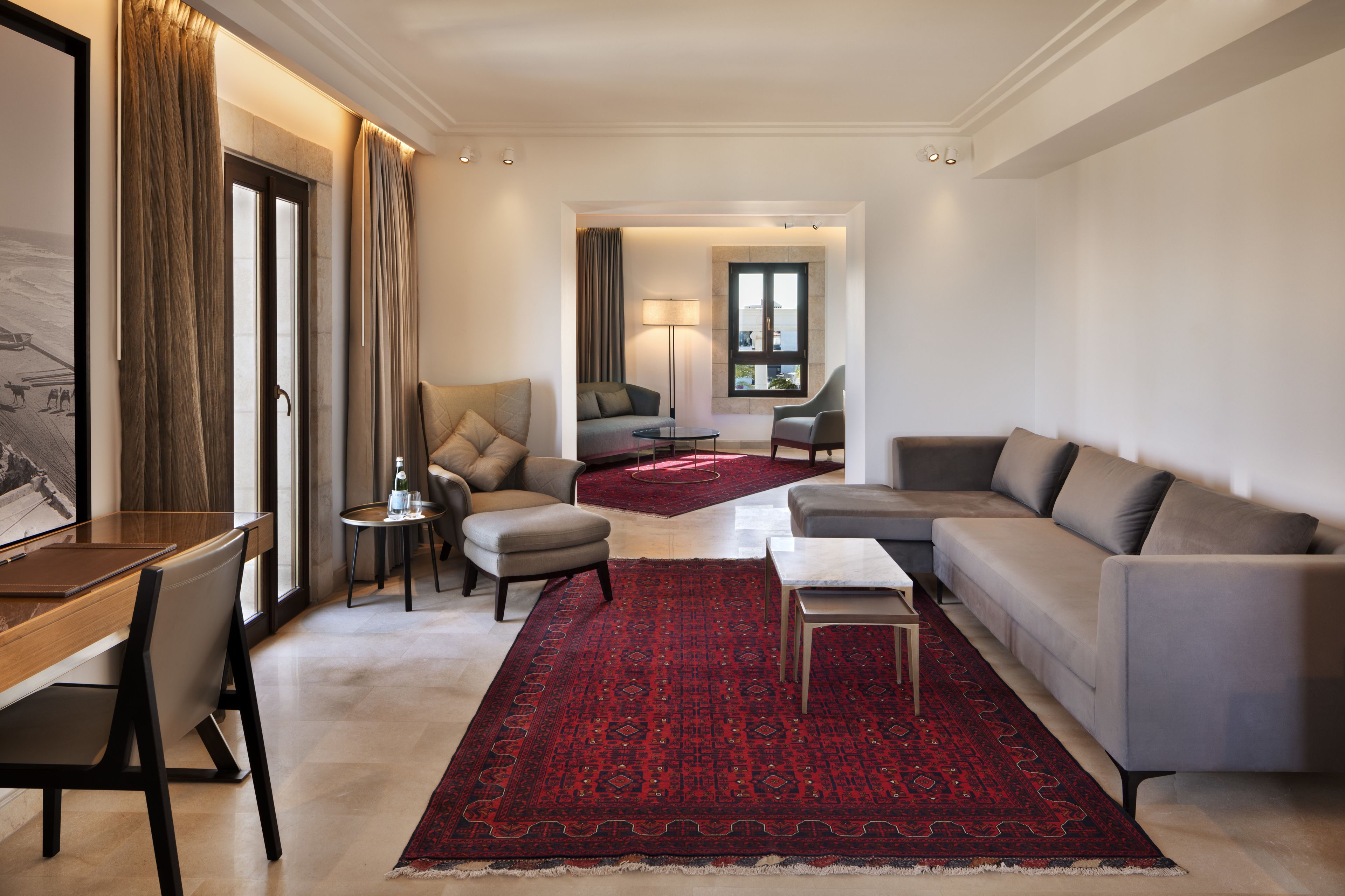 A modern hotel room with a dining area, a sofa, a chair, a small table, and a red patterned rug.