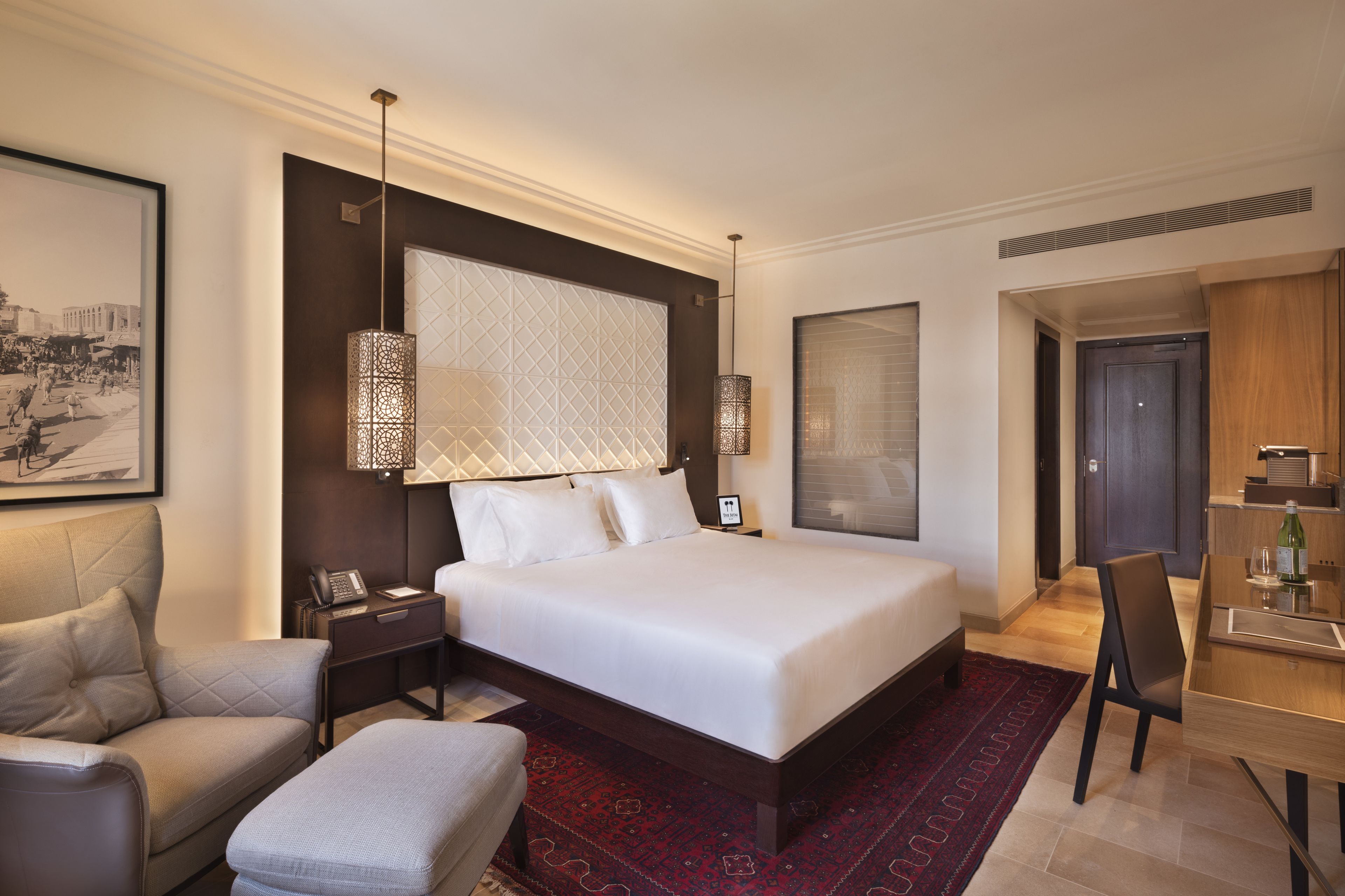 A modern hotel room with a large bed, a desk with a chair, a small sofa, and a red patterned rug.